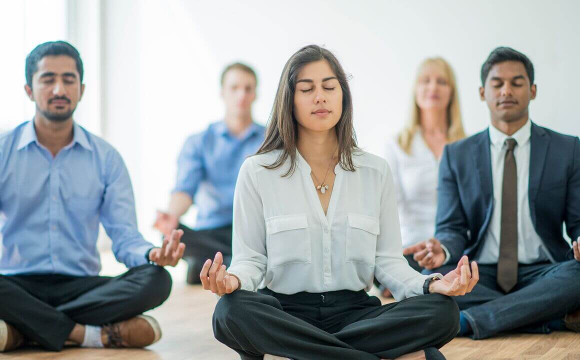 A multi-ethnic group of young business men and women in semi-casual office clothes are sitting on the floor and meditating to relax in an indoor, sunlit office.